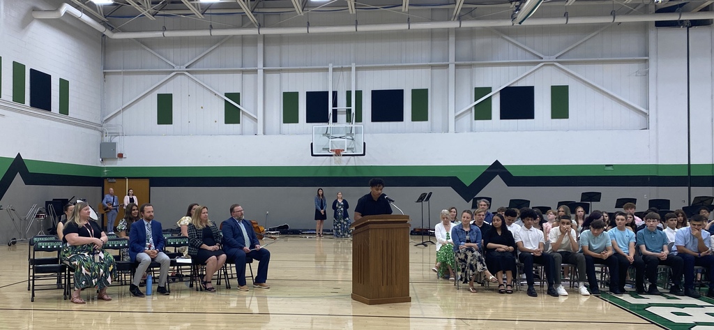 A school event in a gymnasium with a youth speaking at the podium, other adults in seats and students seated and musicians standing in the background.