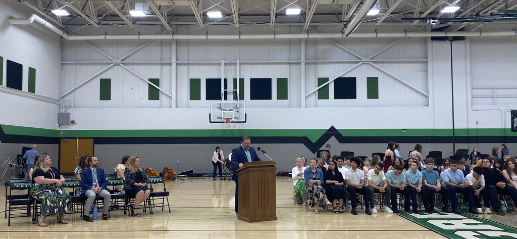 A school event in a gymnasium with an adult speaking at the podium, other adults in seats and students seated and musicians standing in the background.