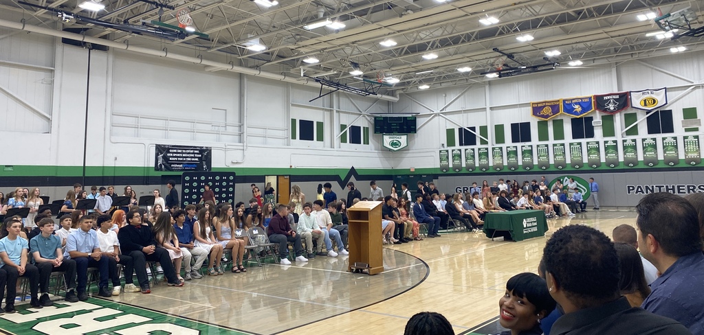 A school event in a gymnasium with students seated and musicians standing in the background.