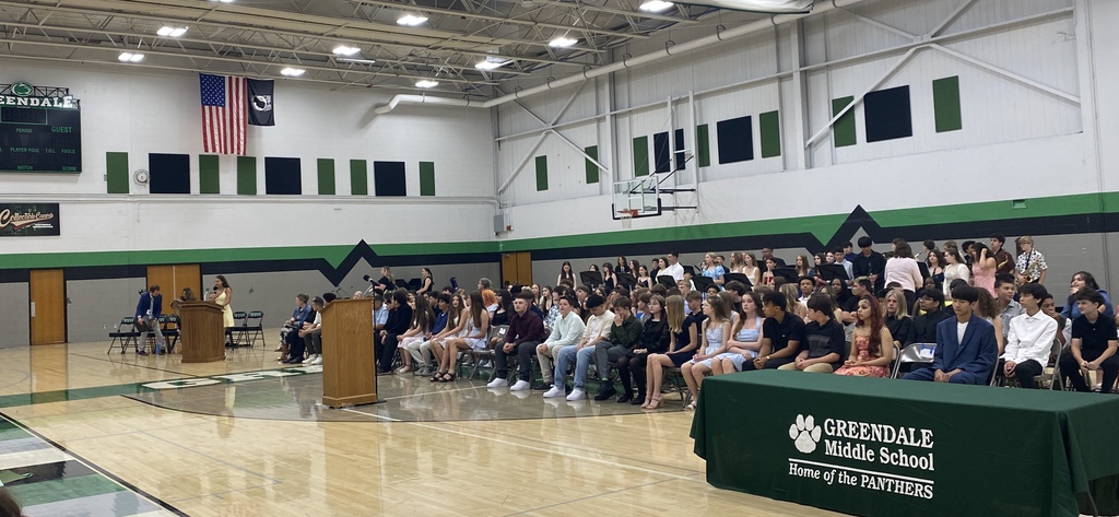 A school event in a gymnasium with a youth speaking at the podium, other adults in seats and students seated and musicians standing in the background.