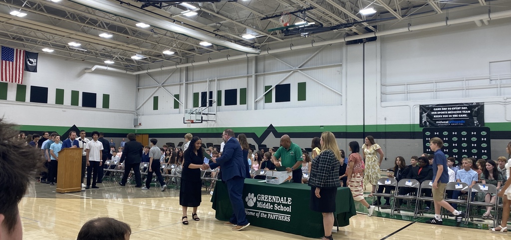 A school event in a gymnasium with students receiving a certificate and shaking hands with adults and other students seated in the background.