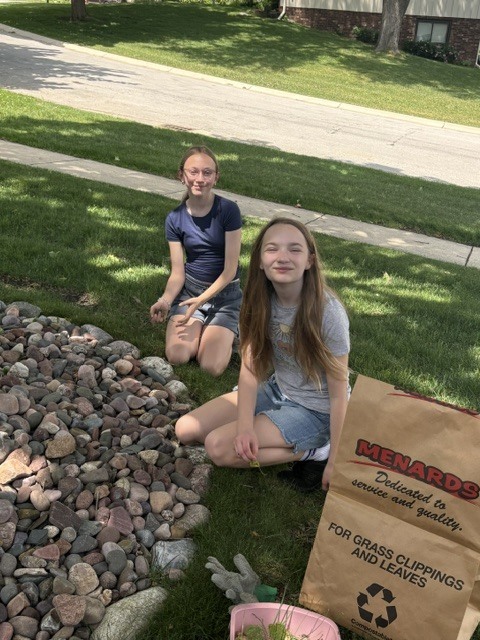 Two students sitting in the grass along a garden pulling weeds.
