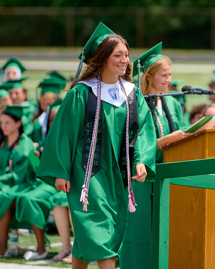 A student wearing a green cap and gown walking in front of a podium.