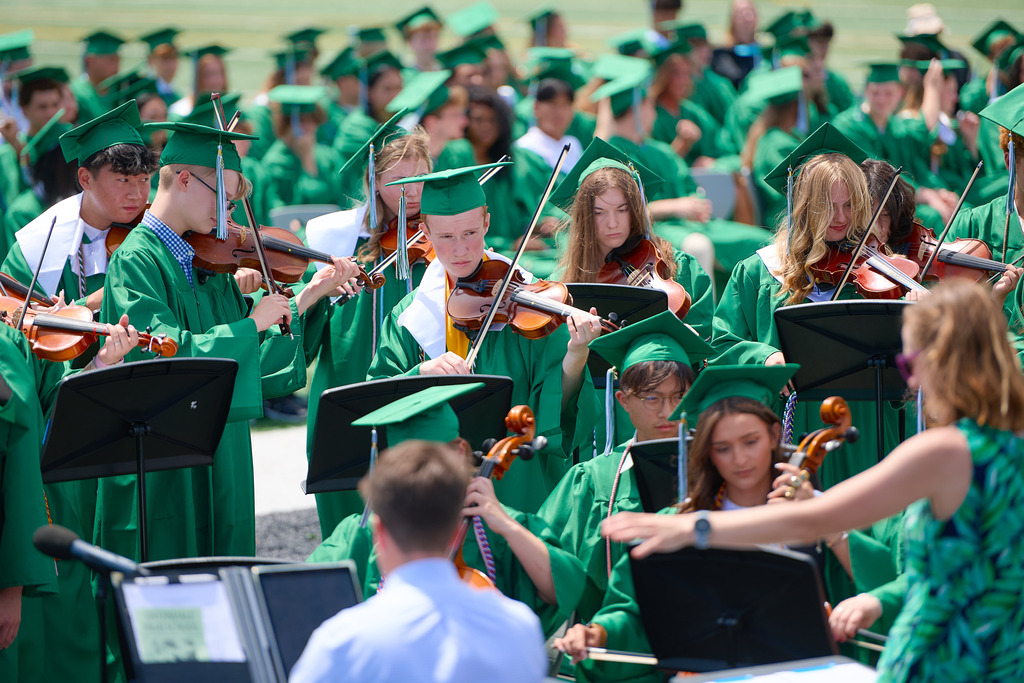 Students wearing green caps and gowns with orchestra instruments performing a song.