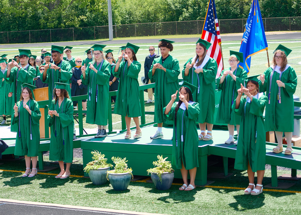 Students wearing Green caps and gowns standing in front of the American flag and signing the National Anthem. 