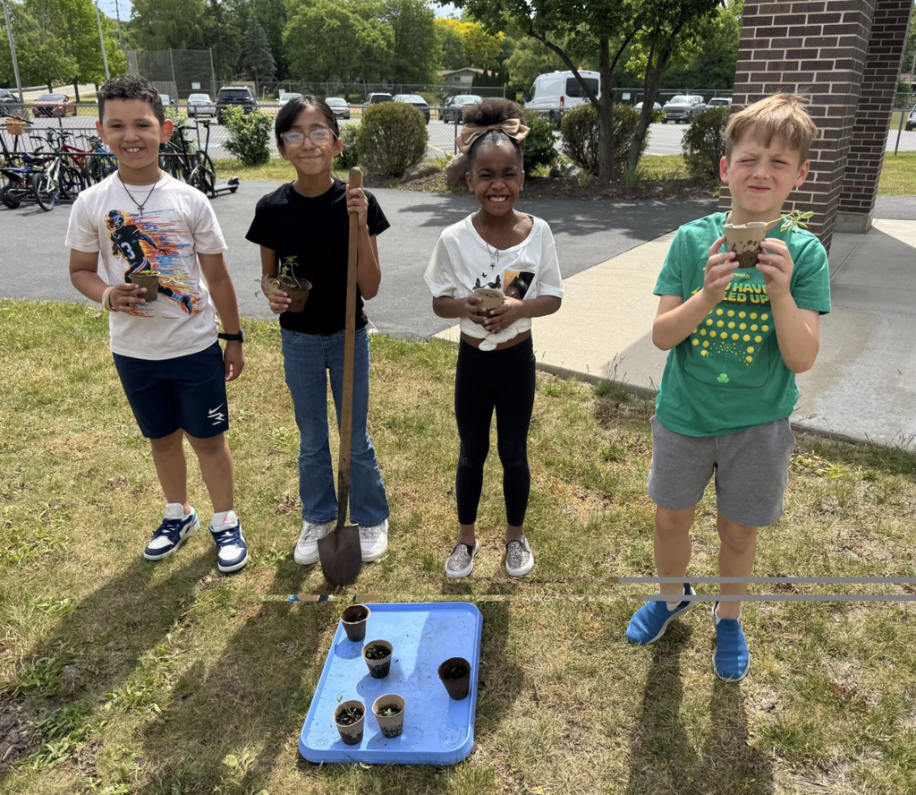 Four students posing in front of the school gardens with garden tools and small plants.