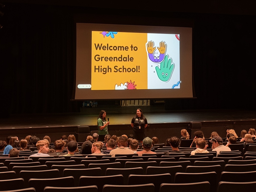 Two teachers standing in front of a group in the auditorium with a screen showing a slide that reads Welcome to Greendale High School.