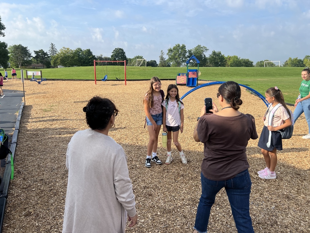 Two students standing and smiling with others looking on while a family member takes their photo.
