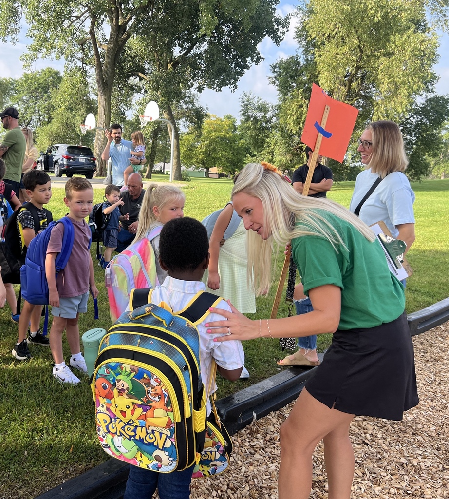 A teacher wearing a green shirt welcoming a student as others look on and line up for the day.