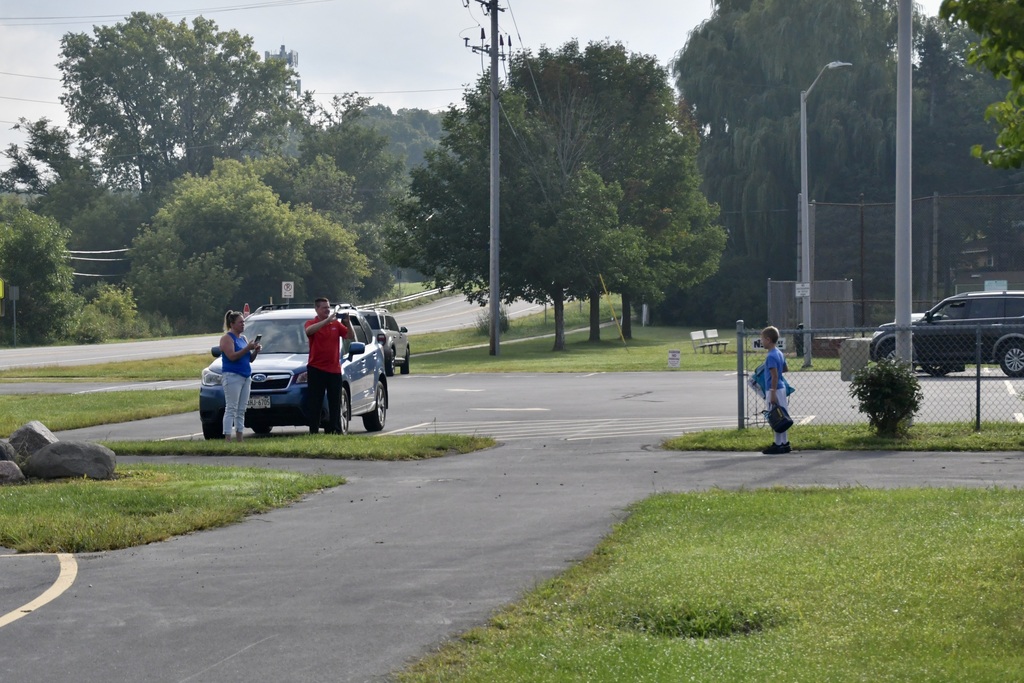 Both parents taking photos with their phones of a boy standing in front of the school with his bags and backpack.