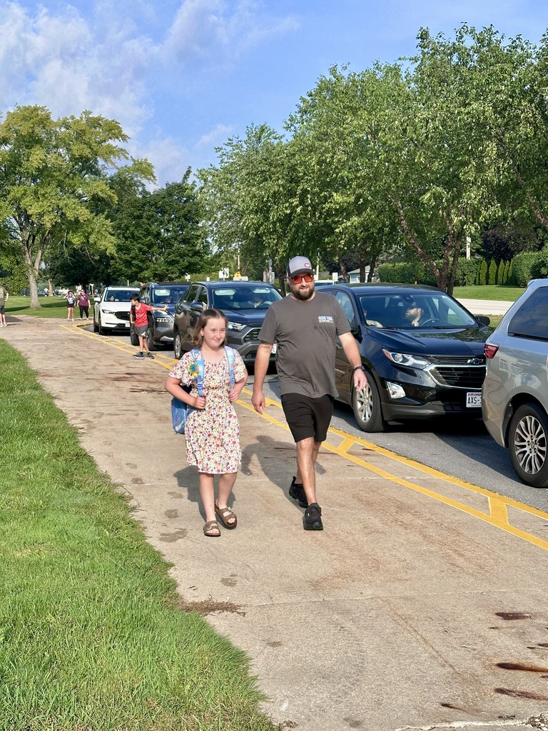 A dad and his daughter with her backpack on walking down the sidewalk in front of the school.