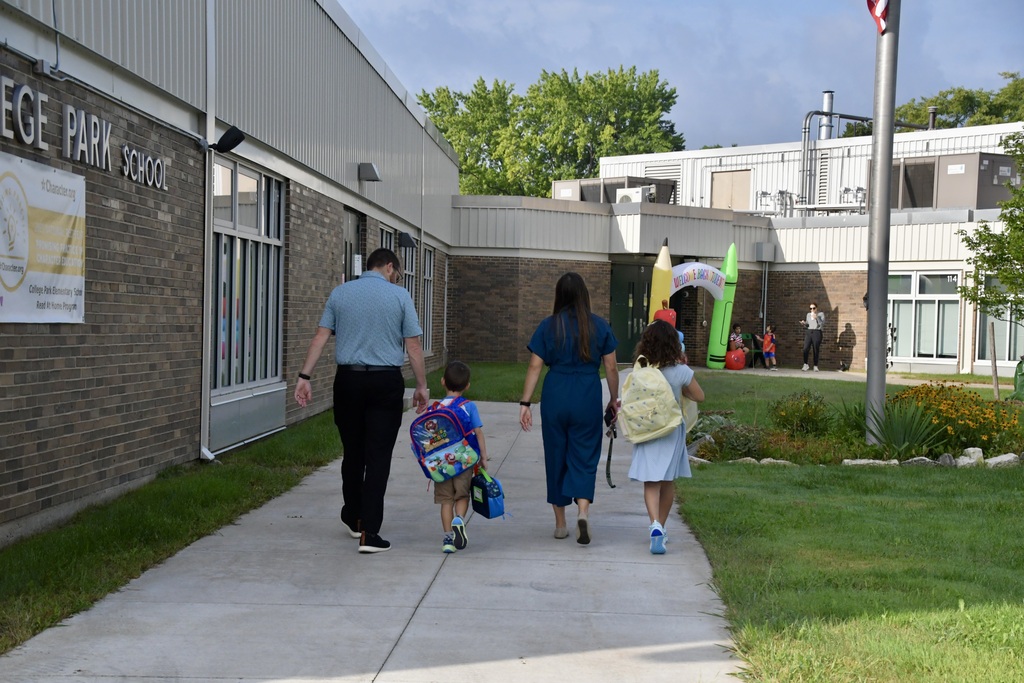 A family (parents and a boy and a girl) from the back walking up the school sidewalk towards the door.