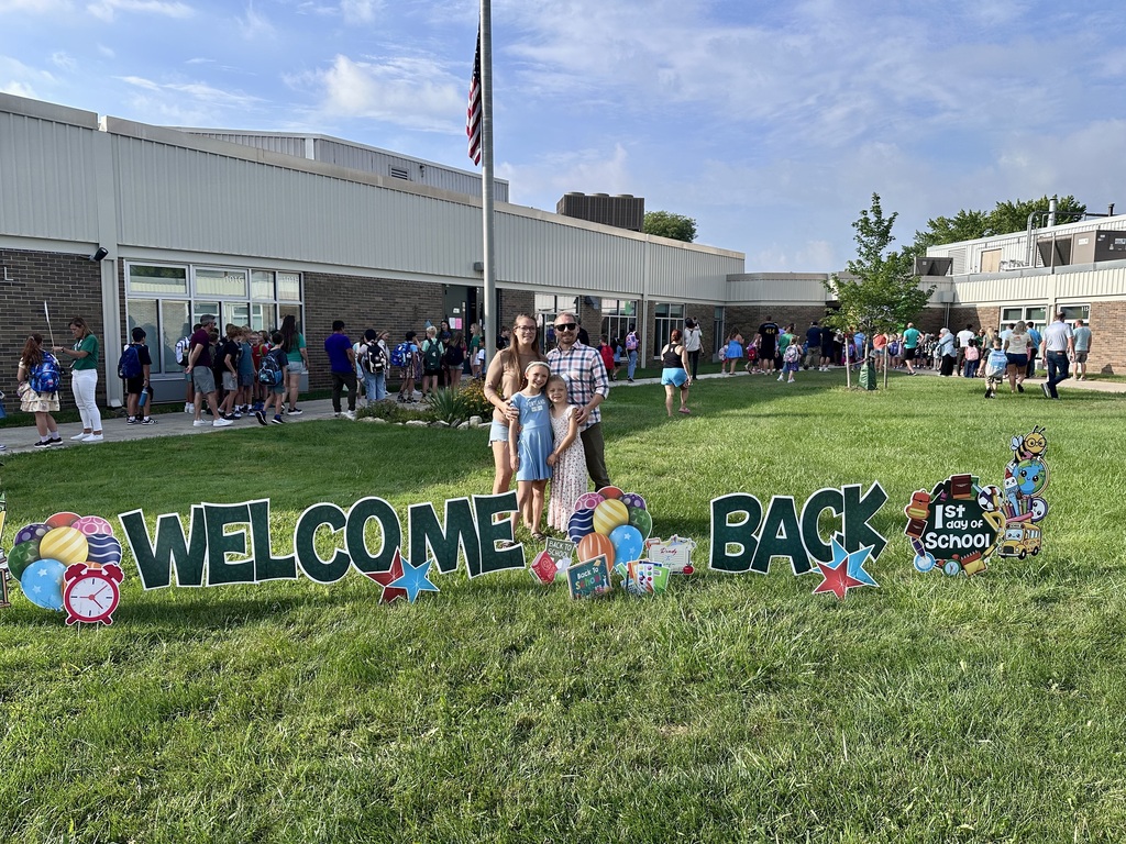 A mom and dad and their two daughters posed in front of the school behind the sign that says "Welcome Back".