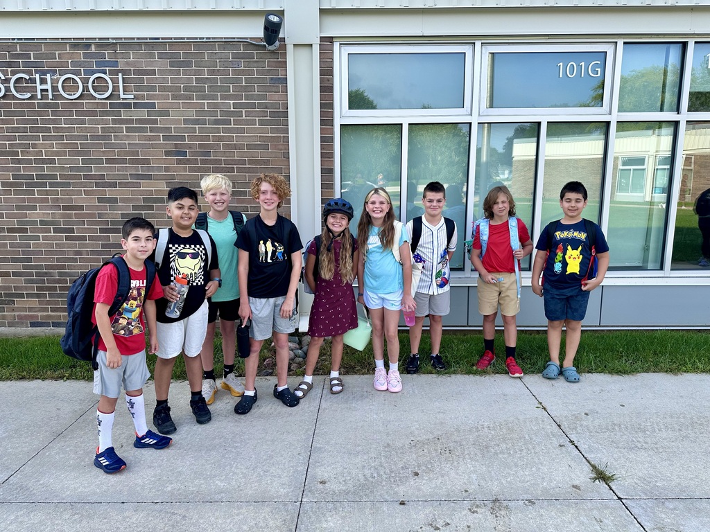 A multi-grade group of boys and girls standing in front of the school, smiling and posing together on the first day of school.
