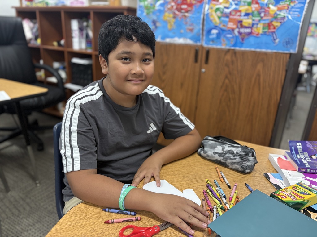 Student smiling at the camera from his desk working with crayons and a scissors.