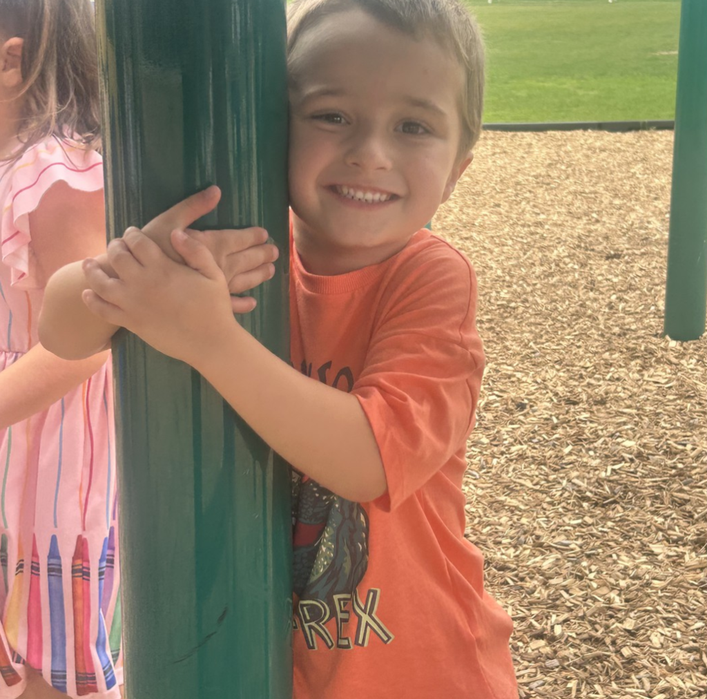 A 5K boy smiling as he hugs a green pole out on the playground.