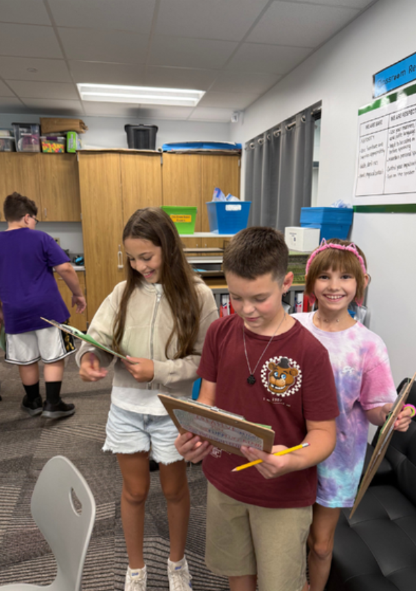 Three fifth graders are smiling and holding clip boards as they conduct a classroom scavenger hunt.