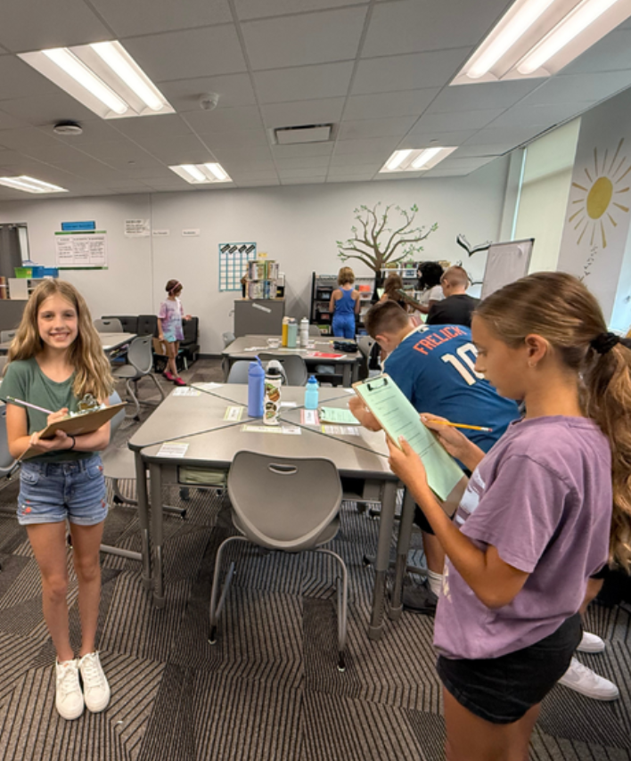 Two fifth graders are holding clip boards and working on a classroom scavenger hunt.