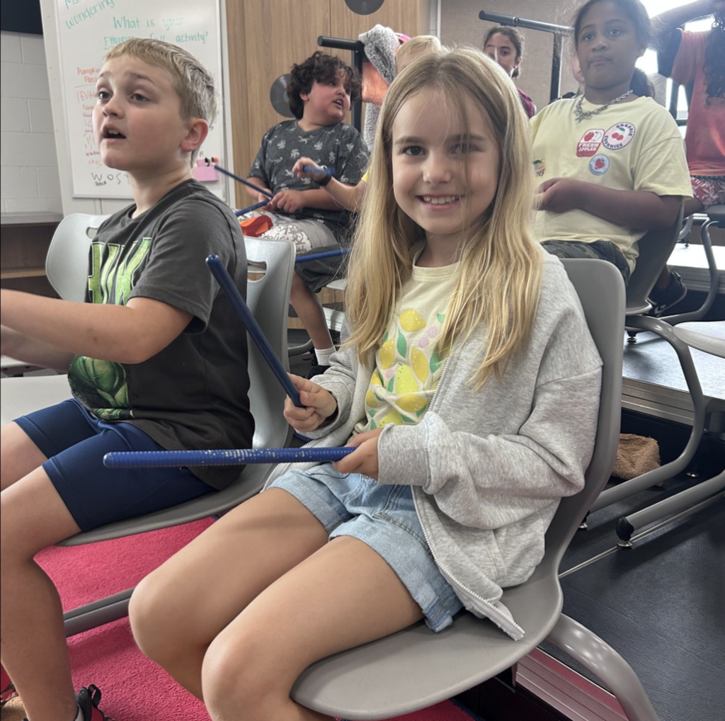A smilingirl sitting among other students using wooden sticks to learn about music.