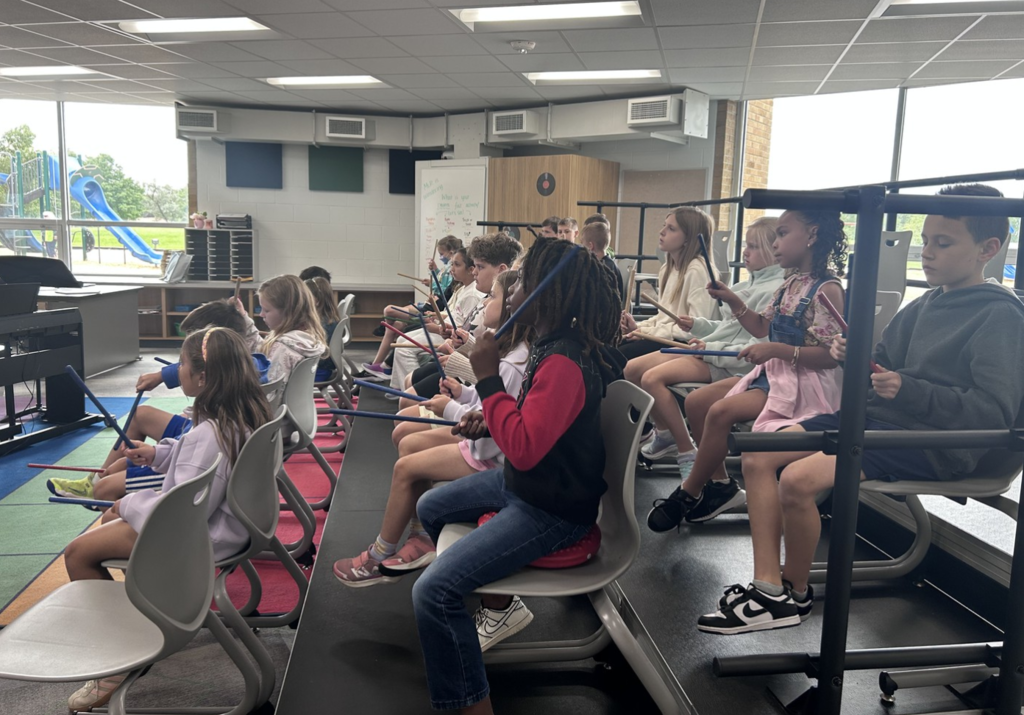 A sideview of students sitting in riser seats using wooden sticks to learn about music.