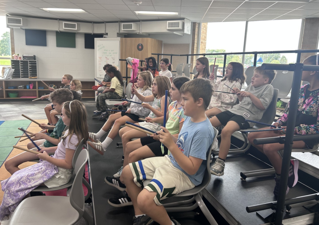 A side view of students sitting in riser seats facing the teacher using wooden sticks to learn about music.