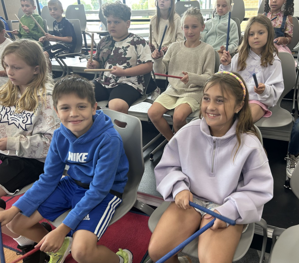 Two smiling students with others around them using wooden sticks to learn about music.