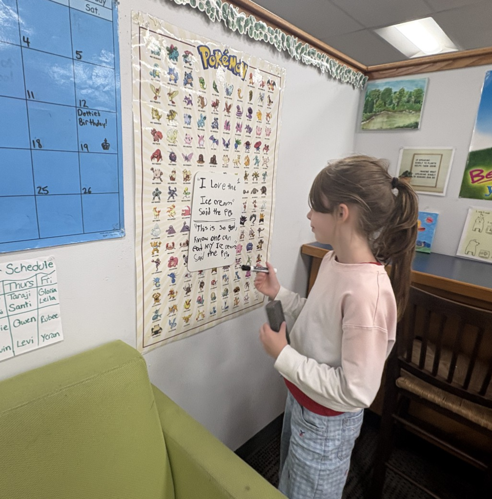 Fourth grade student stands by small white board on the wall with a marker in her hand looking at work on the board.
