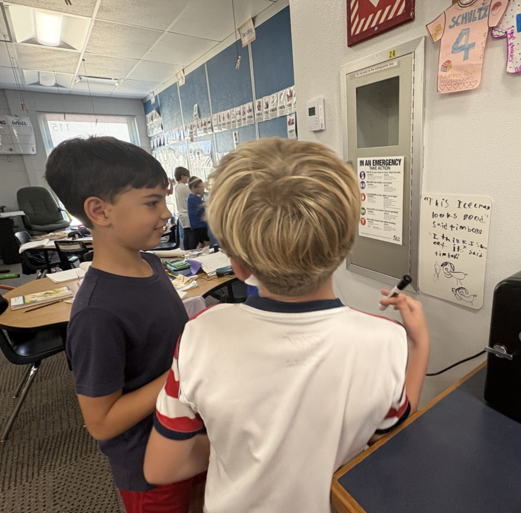 Two fourth graders conferring while looking at notes on a small white board on the wall.