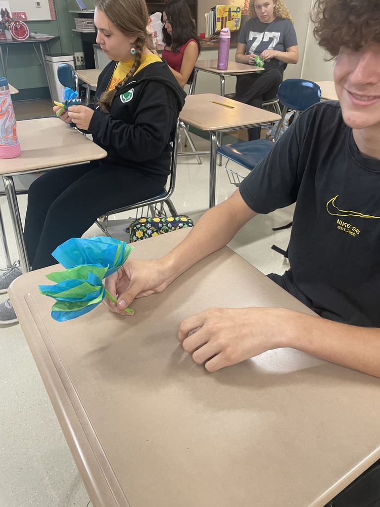 One smiling student holding a paper flower with others in the background working on their flowers.