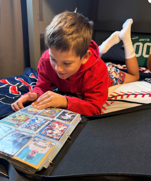 A boy laying on his bed looking through his baseball card collection in a binder.