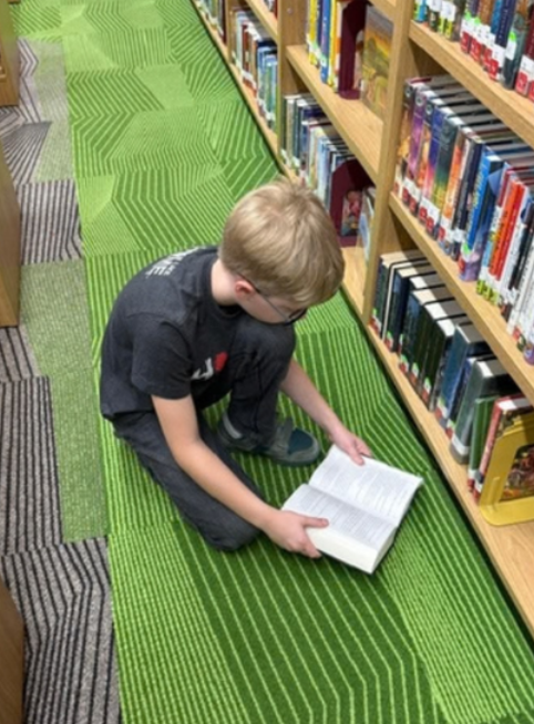 A boy sitting on the floor at the school library looking at a book.