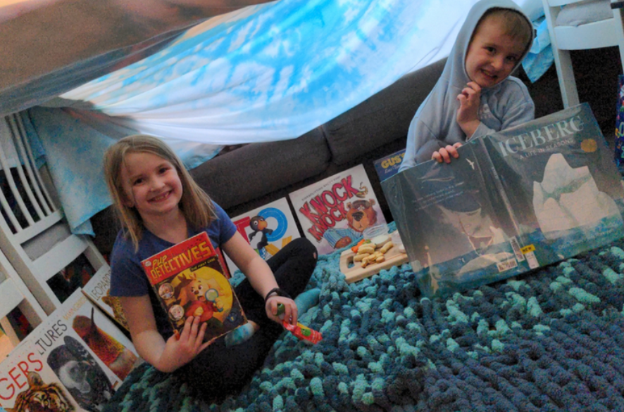 Siblings enjoy some fun reading time under a homemade blanket fort.
