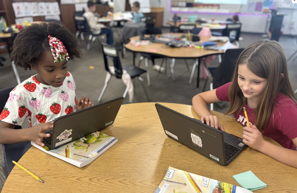 Two girls at a table with laptops confer as they share their writing.