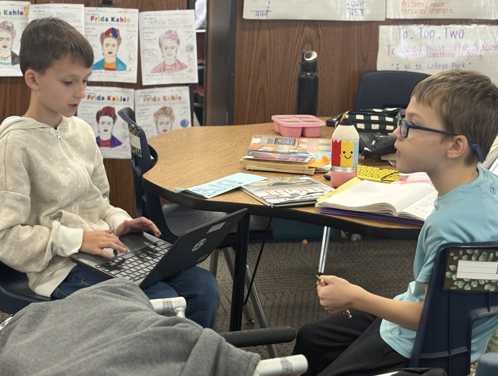 Two boys sit together at a table, one with a laptop, as they share their writing.