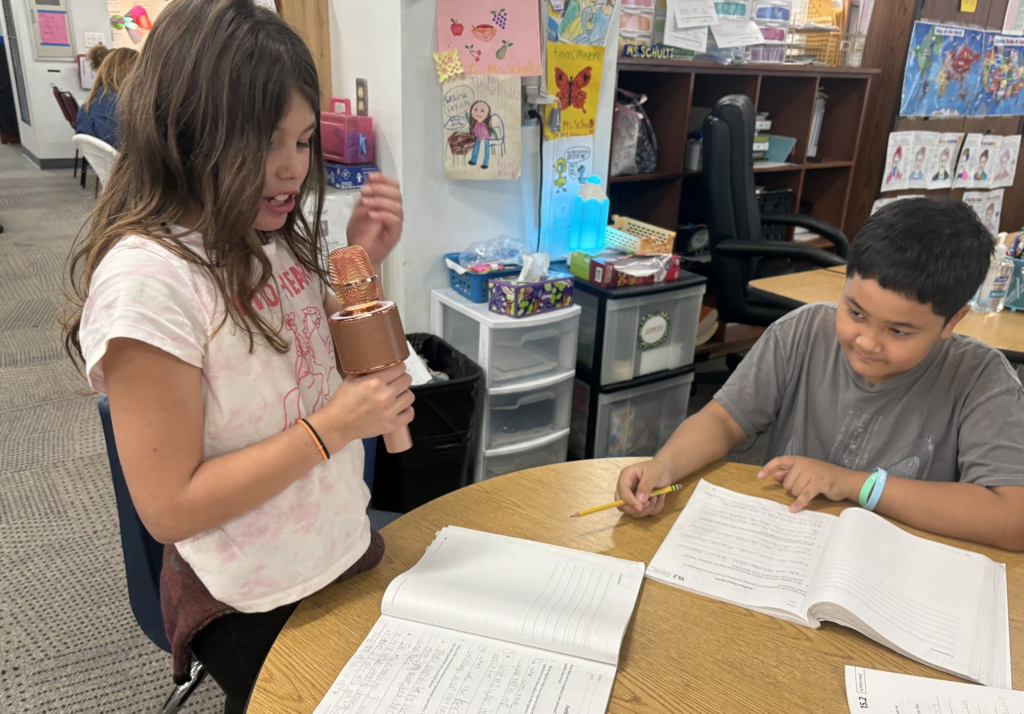 A girl and a boy sit together at a table with workbooks in front of them as they share their writing.