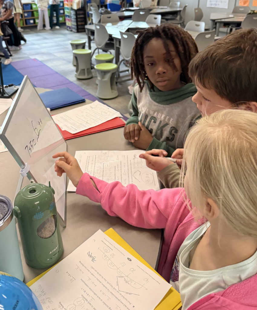 Three students confer at a desk with papers in front of them and one student pointing to an equation on a small whiteboard.
