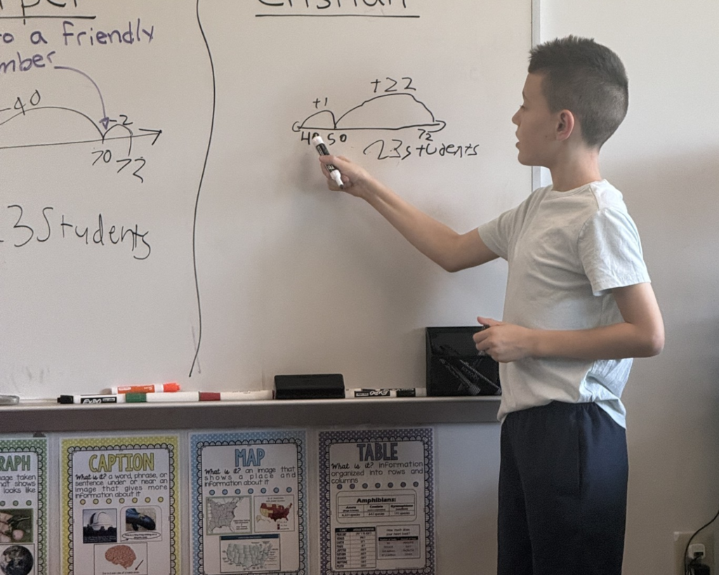 One student stands at a big whiteboard holding a marker and explaining the equation on the board.
