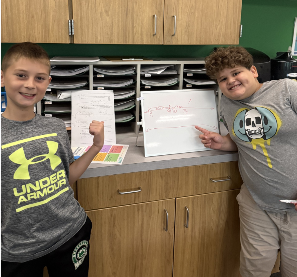 Two students smile and point to their Math work which is displayed on boards standing on a counter behind them.