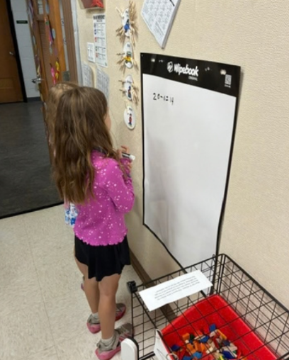 A student shown from the side holding a marker and working on a math equation on a whiteboard.