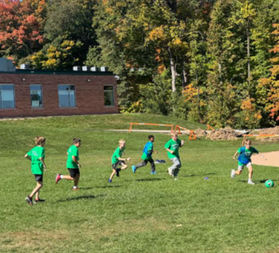 Five students running after another student kicking a ball on a grassy field playing speedball.