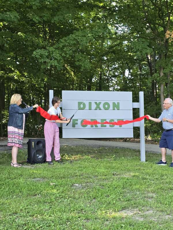 Three people standing in front of a sign cutting a red ribbon with a big scissors