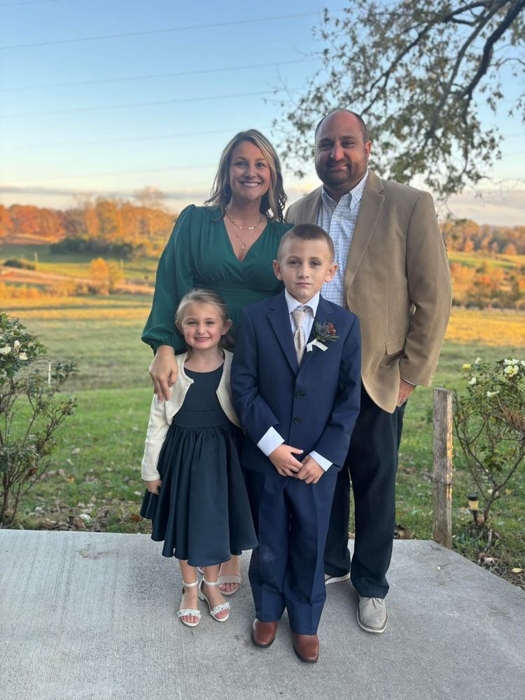 A family of four stands together outdoors on a concrete patio with a scenic, sunlit countryside landscape in the background. An adult woman in a deep green dress stands beside an adult man wearing a tan blazer, blue shirt, and dark pants. In front of them, a young girl in a navy dress and white cardigan stands next to a young boy dressed in a dark blue suit with a boutonniere. Trees with autumn foliage and rolling fields stretch out behind them under a clear sky.