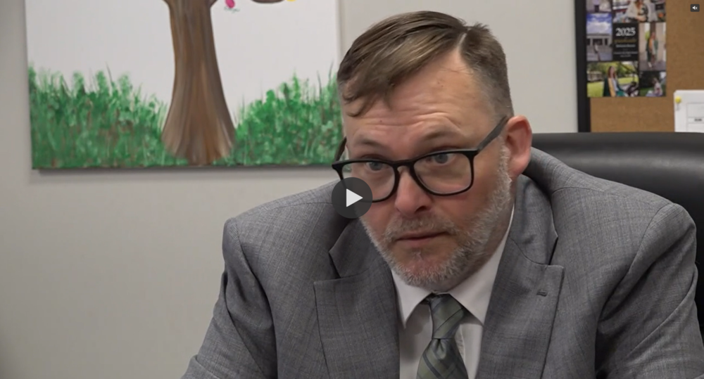 Dr. Jason Horne, Assistant Director of Greeneville City Schools, seated in an office wearing a gray suit, white shirt, and patterned tie. Behind him is a painting of a tree with green foliage and a corkboard with various pinned papers and images