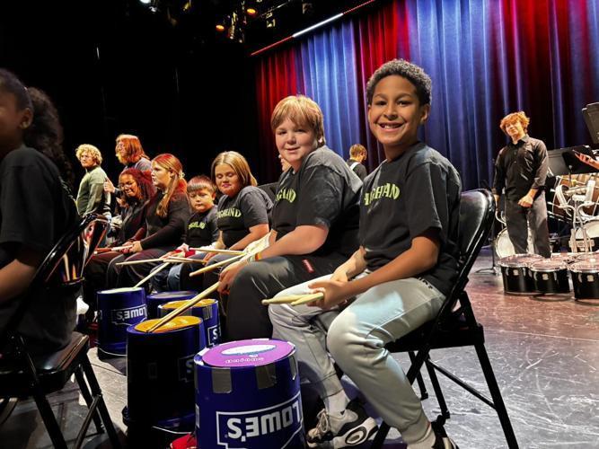 A group of students sits in folding chairs on a stage holding drumsticks, each playing on brightly colored practice buckets labeled with store branding. They wear matching dark T‑shirts, and stage lights cast warm colors across the curtains behind them. Additional students and percussion equipment are visible in the background, creating an energetic pre‑performance atmosphere.