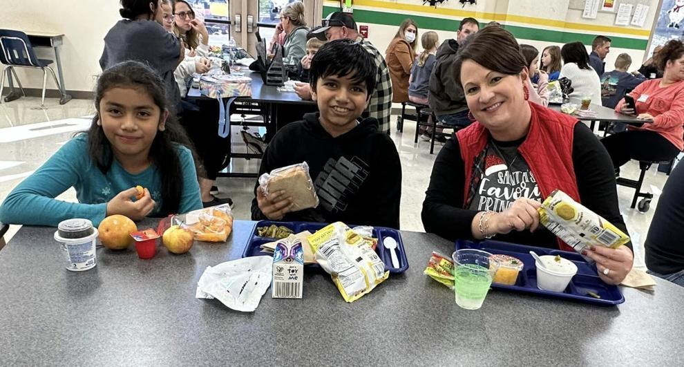 Three people sitting at a cafeteria table eating lunch, with various food items like yogurt, fruit, a sandwich, milk, chips, and salad on their trays. More people are visible in the background, also seated and eating.