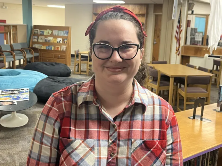A person stands indoors in what appears to be a library or media center, wearing a red and blue plaid shirt and a red headband. Behind them are wooden bookshelves, tables, chairs, and beanbag seating, along with an American flag and various books and materials displayed around the room.