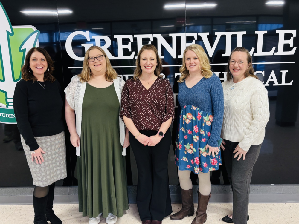 A photo from the celebration shows five smiling women in business casual attire standing in front of a wall that reads “GREENVILLE” with a partially visible green student logo.