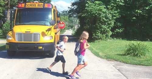 “Two children cross the street in front of a stopped yellow school bus with its stop sign extended, surrounded by green trees and grass.”