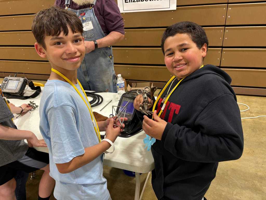 two elementary school boys hold welded metal objects in front of a table with various welding implements