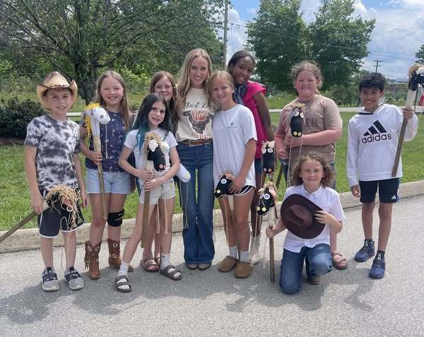 **Alt Text:** A group of ten children and one adult stand outdoors on a paved area with grass and trees in the background. The children are holding hobby horses, and one child is kneeling in front holding a hat. The adult stands in the center wearing a t-shirt and jeans. The sky is partly cloudy with patches of blue.